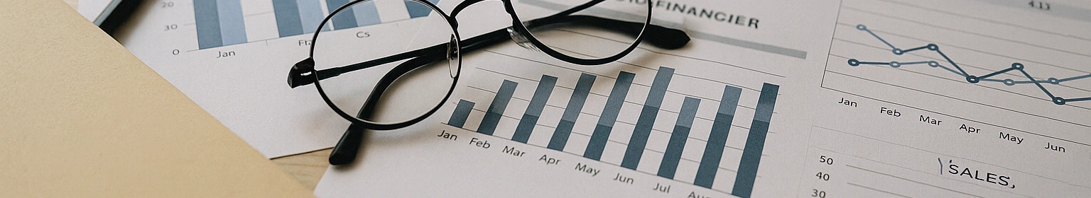 Documents financiers imprimés, lunettes et stylo posés sur une table, illustrant une pause utile pour analyser et ajuster le pilotage avant la rentrée.